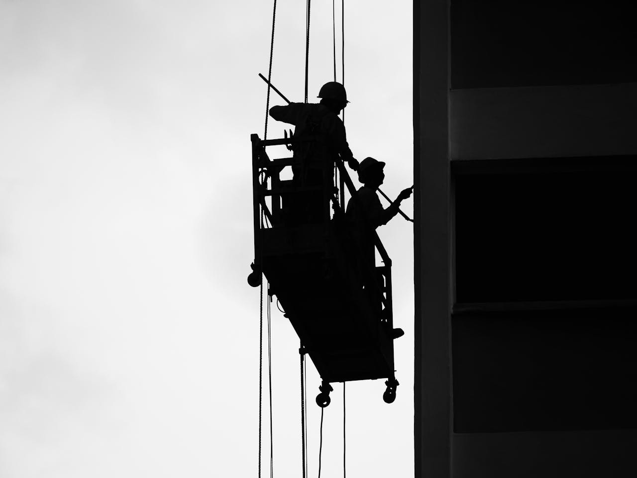 Anasayfa Two workers silhouetted on a suspended scaffold against a building facade.